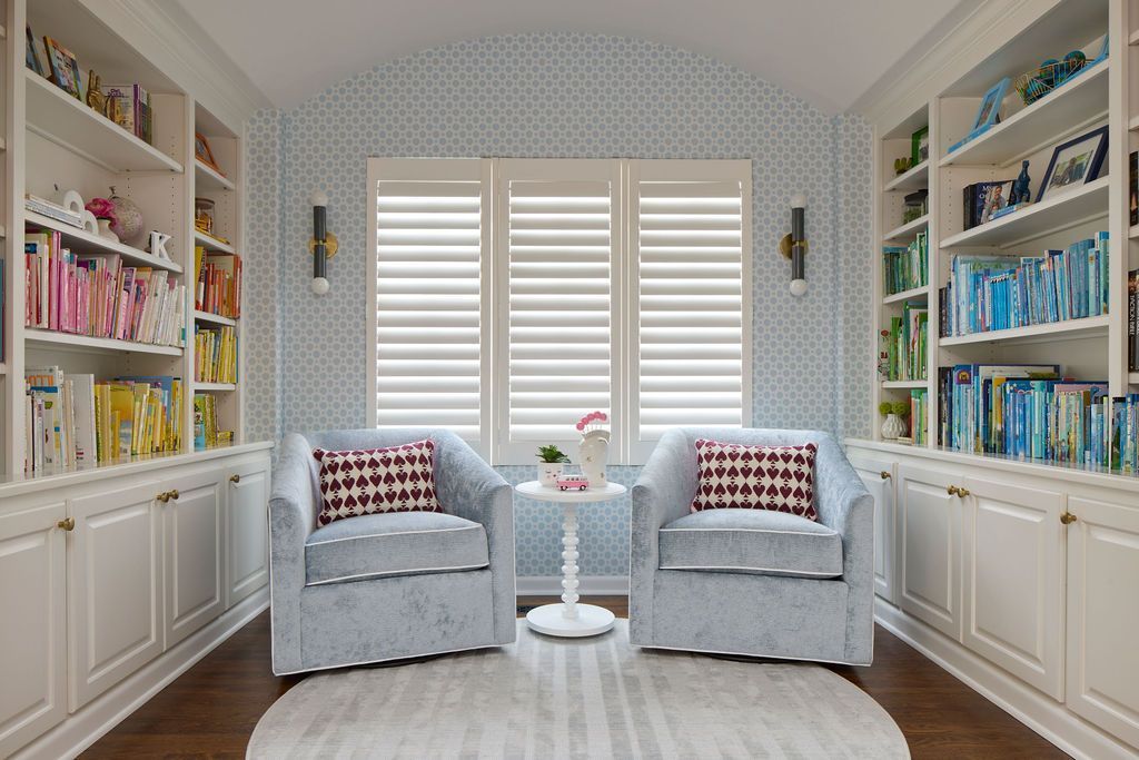 Cozy reading room with built-in bookshelves, two blue armchairs, round rug, and window with shutters.