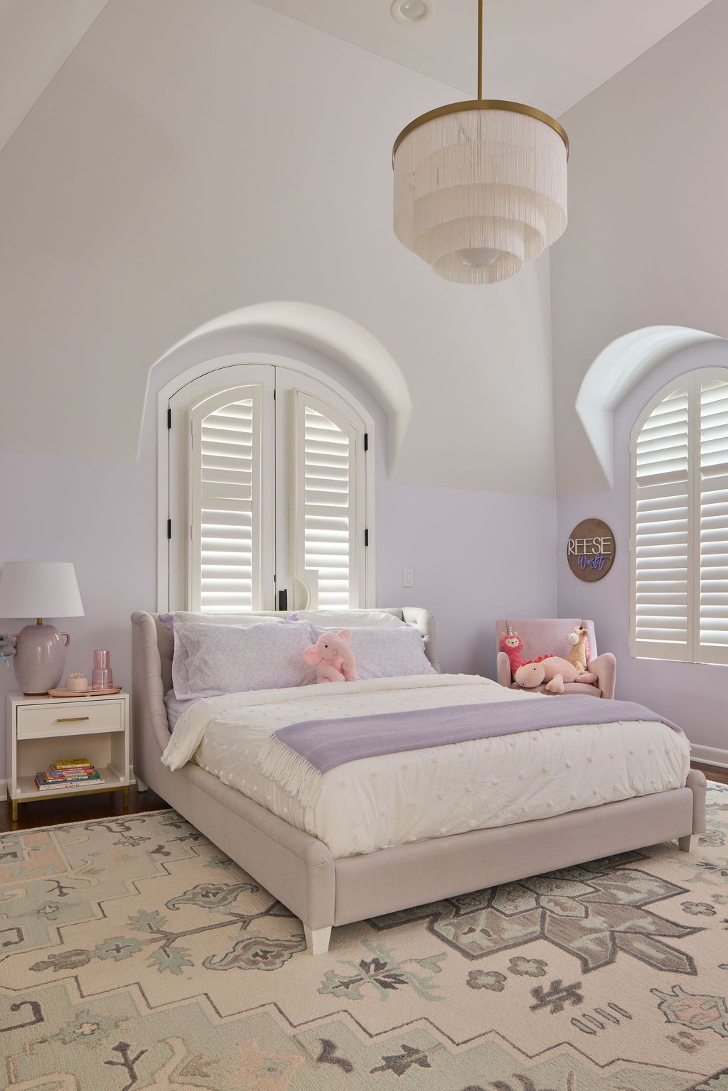 Bedroom with light purple walls, bed, rug, and arched windows. Gold chandelier hangs from the ceiling.