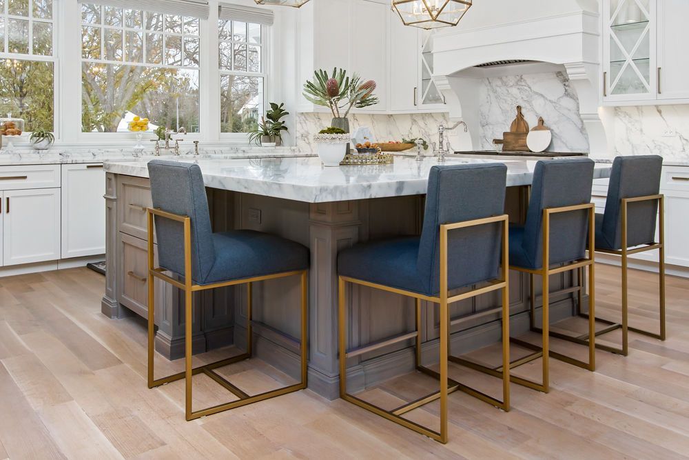 Kitchen island with blue upholstered bar stools, gold frames, white marble countertop.
