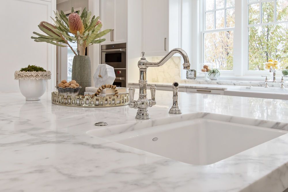 Marble countertop with built-in white sink and chrome faucet. Kitchen with window and floral arrangement.