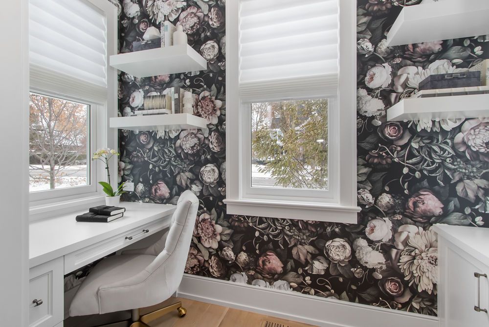 Cozy home office with floral wallpaper, desk, and white chair. Windows offer outside view.