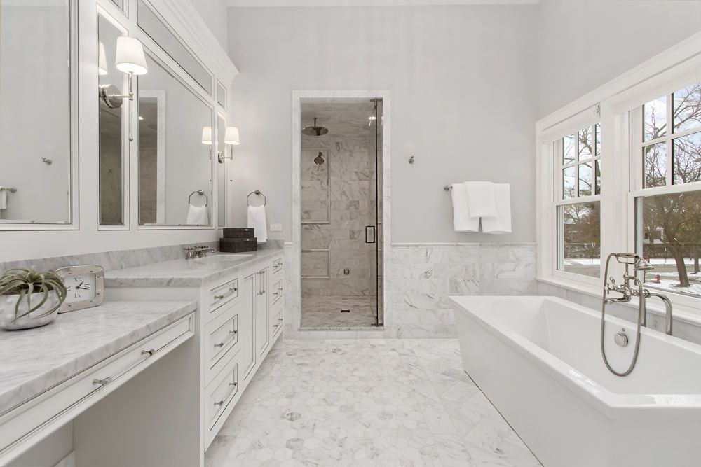 Bright white bathroom with marble counters, vanity, tub, and walk-in shower. Window view of outdoors.