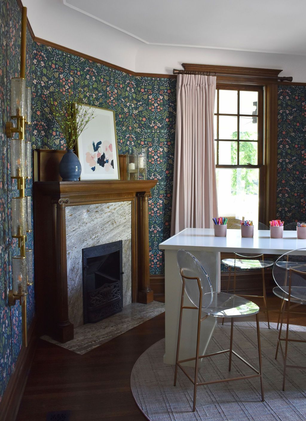 Room with floral wallpaper, fireplace, white table, ghost chairs, and pink curtains.
