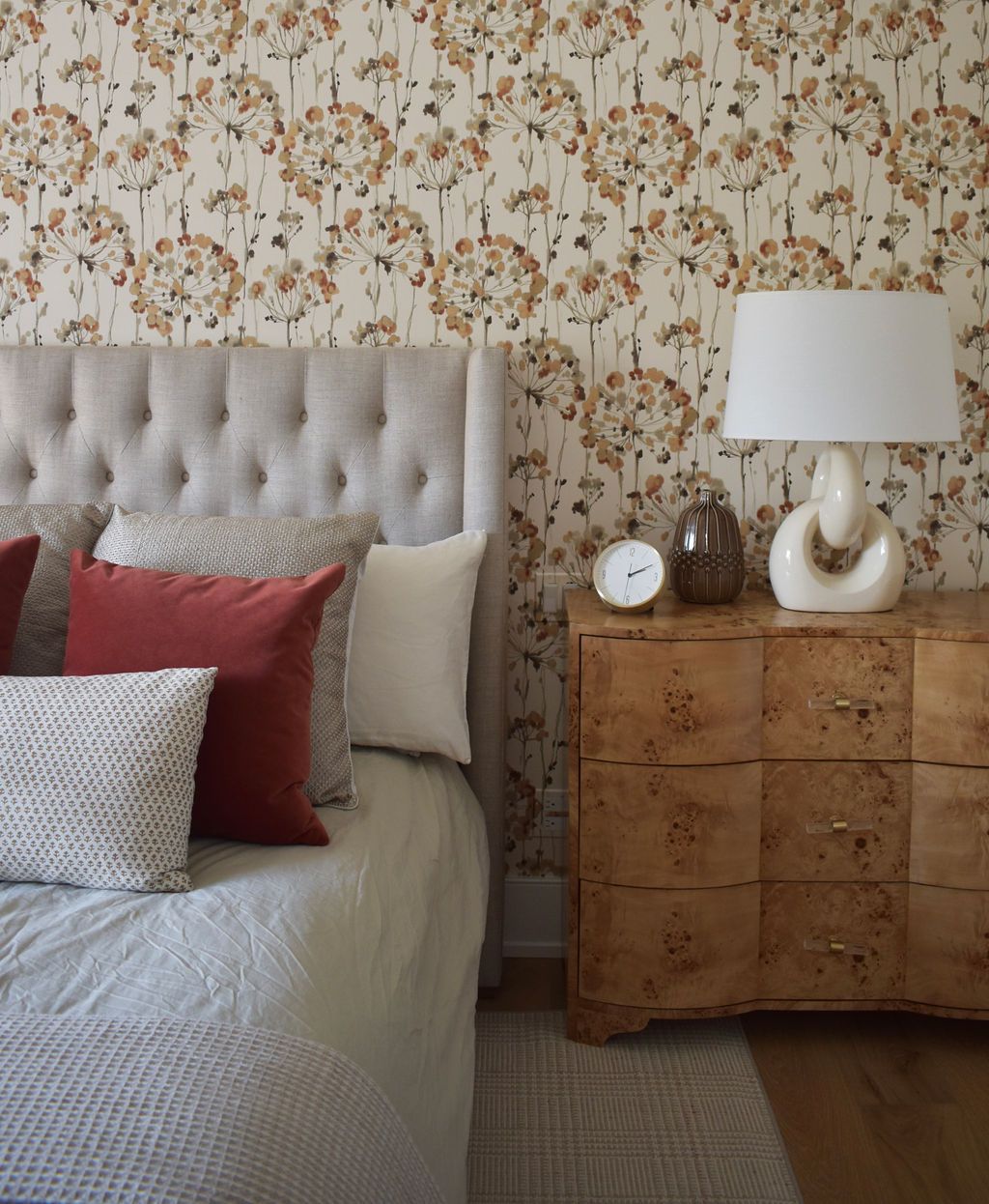 Bedroom with floral wallpaper, tufted headboard, and wooden nightstand with lamp and decor.