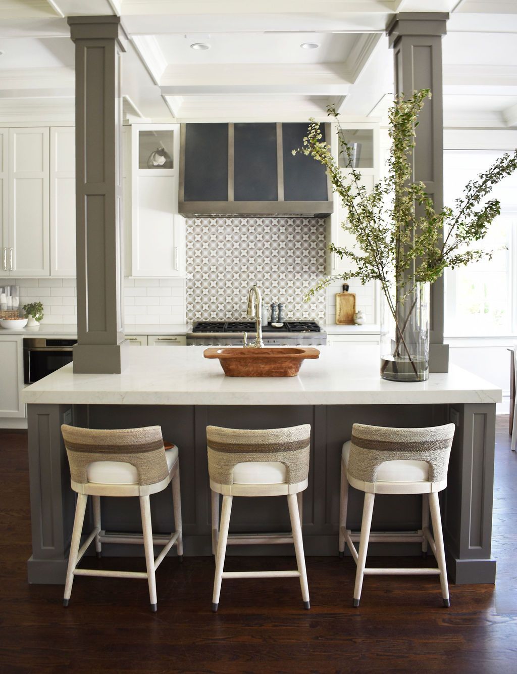 Kitchen island with three stools. Gray cabinets, white countertops, and a vase of branches.