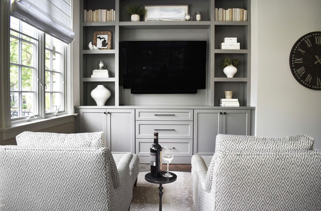 Gray built-in media center with a TV, flanked by bookshelves. Two patterned armchairs face the center, with a small table.