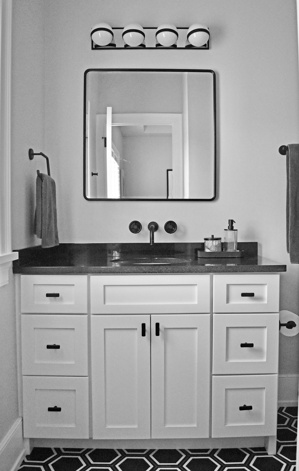Bathroom vanity with square mirror, light fixture, black countertop, white cabinets, and hexagonal tile floor.
