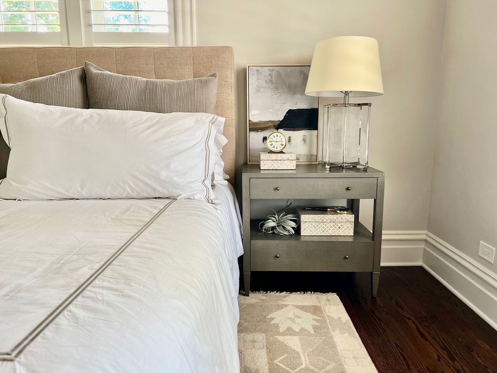 Bedroom with bed, nightstand, lamp, and artwork. Neutral tones, hardwood floor, and a small rug.