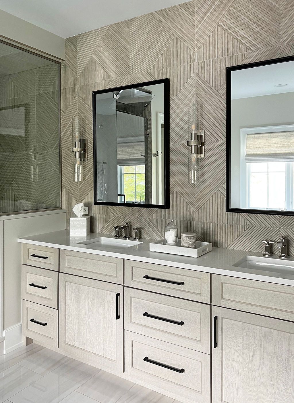 Bathroom with light gray double vanity, black-framed mirrors, patterned beige tile, and sconces.