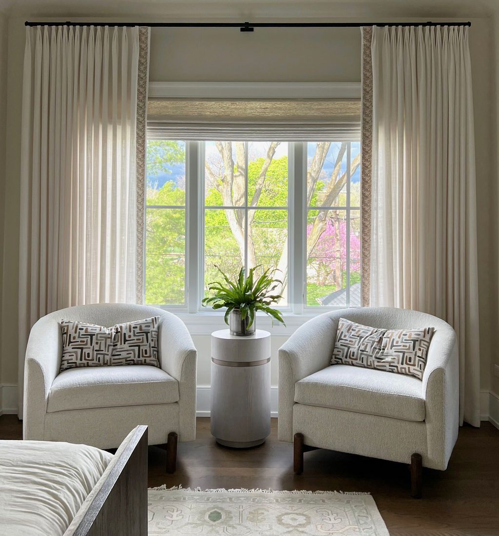 Two white armchairs face a window, flanked by cream curtains. A small table holds flowers.