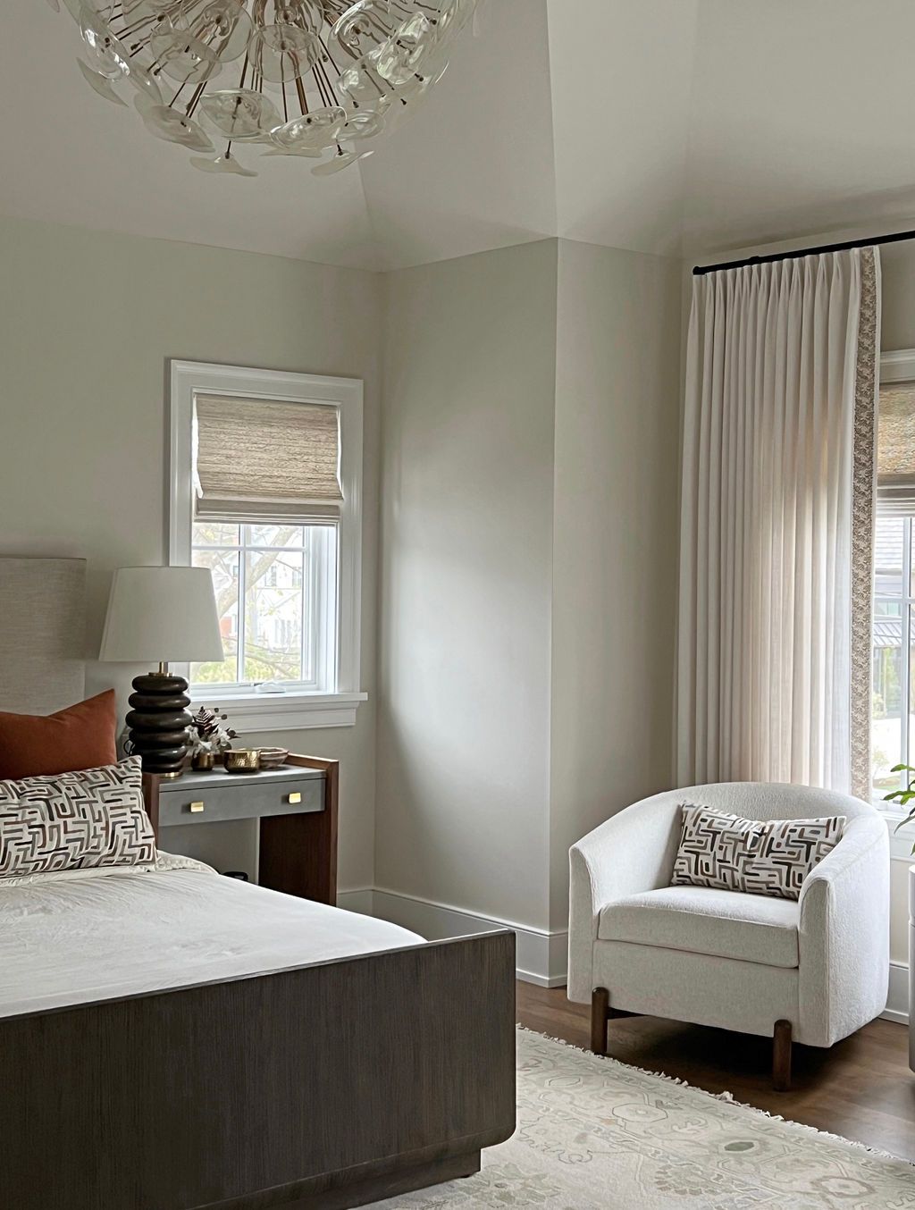 Bedroom with a bed, armchair, window with shades and curtains, neutral color palette.