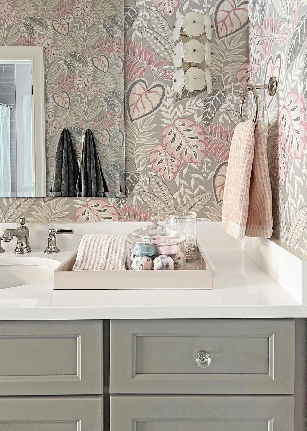 Bathroom vanity with floral wallpaper, towels, and decorative items. Gray cabinets and white countertop.