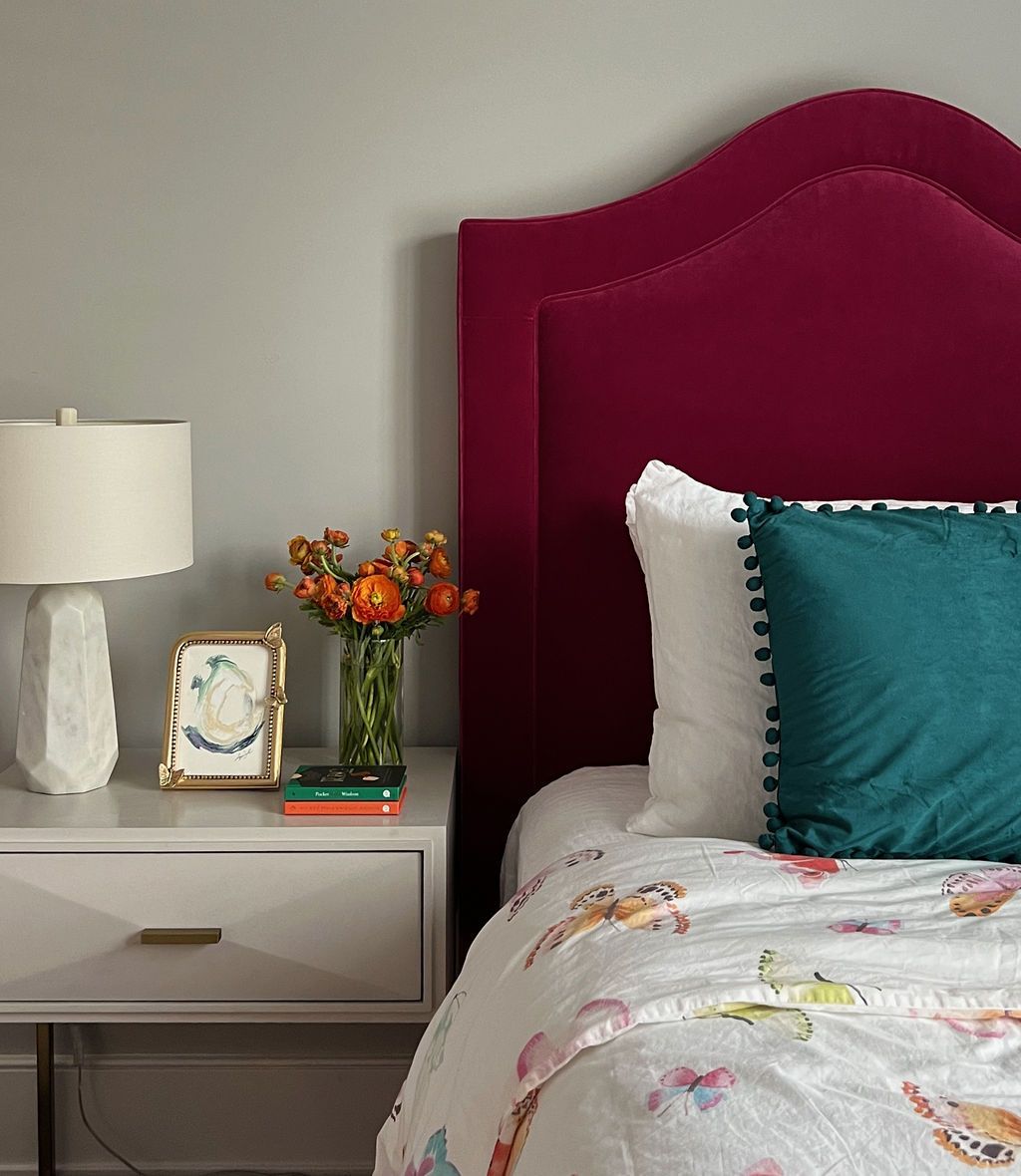Bedroom scene: pink headboard, gray nightstand, teal pillow, floral bedding, flowers, and lamp.