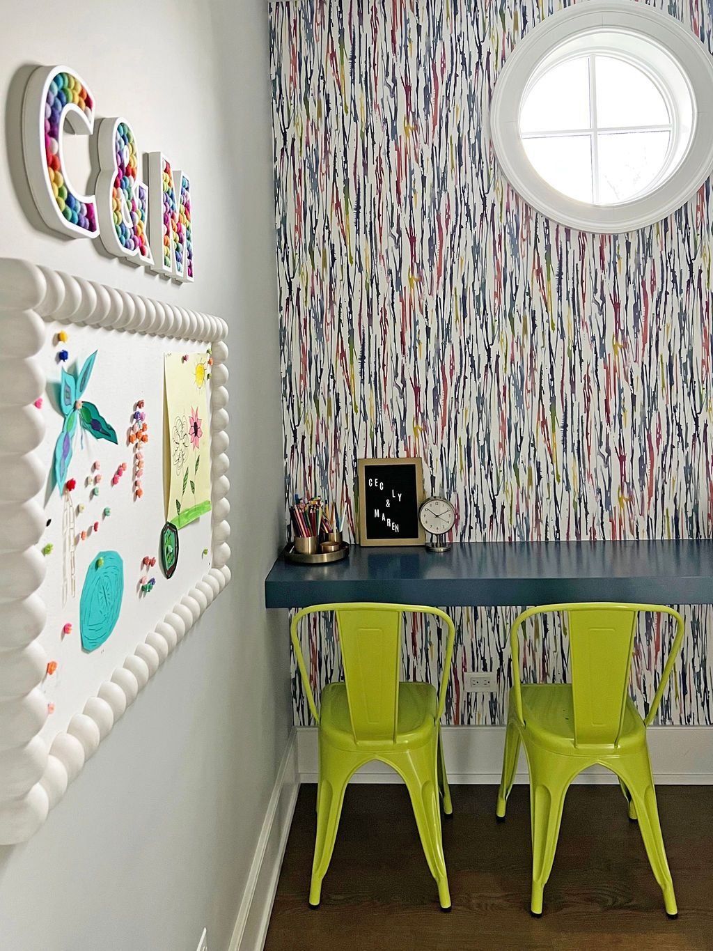 A children's study area with a desk, two green chairs, colorful wallpaper, and a bulletin board with artwork.