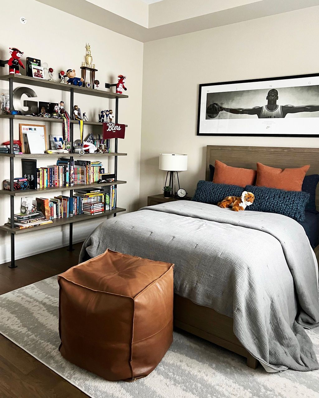 Bedroom with gray bedspread, brown ottoman, wooden shelves filled with items, and a large framed photo.