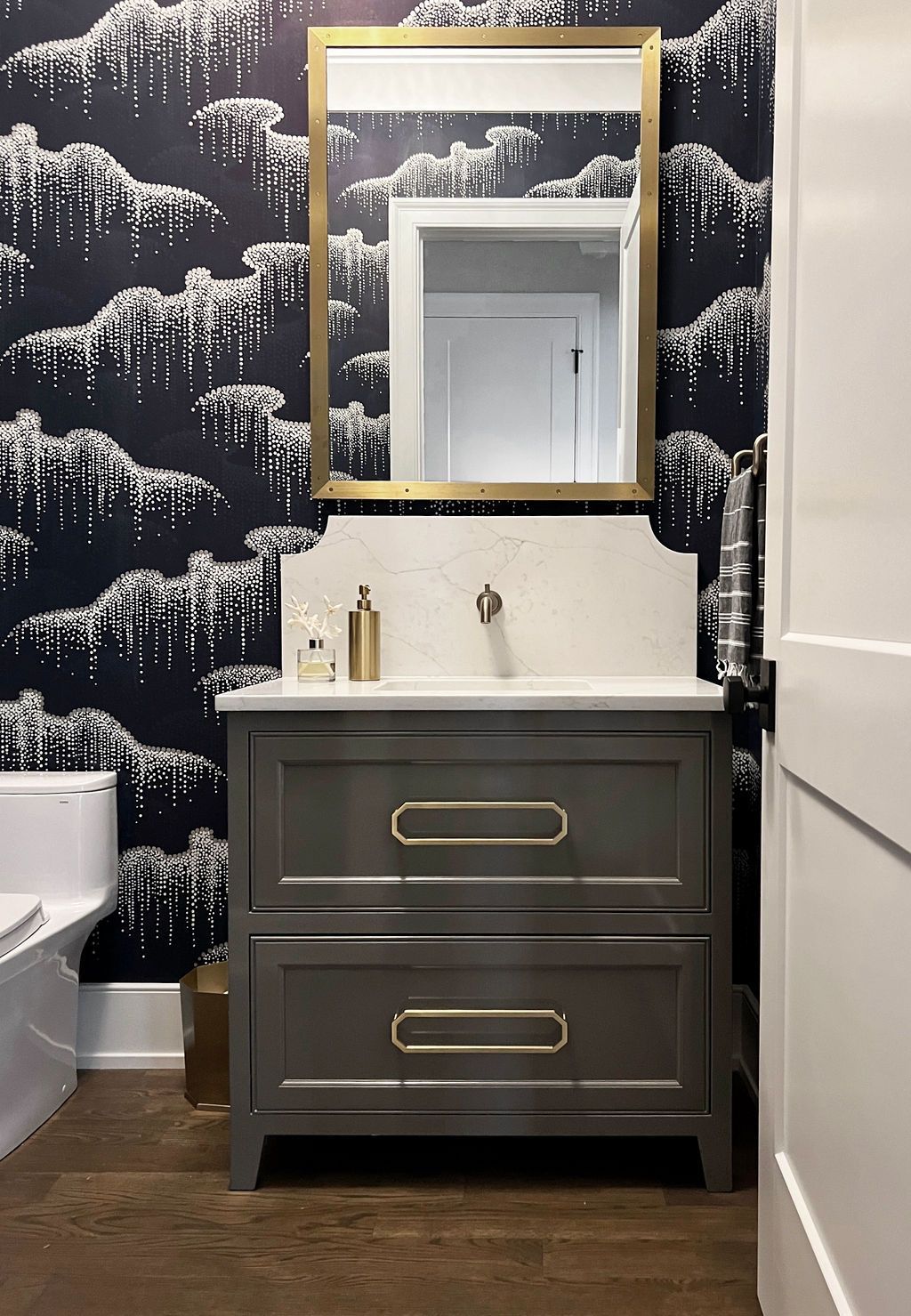 Gray vanity with gold hardware and mirror in a bathroom with patterned wallpaper.