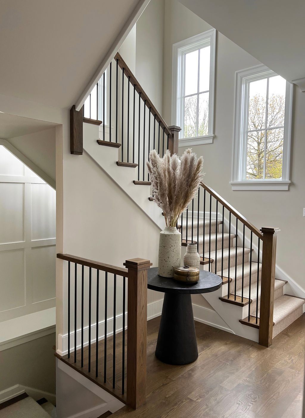 Staircase with wooden handrails and black metal balusters; vase of pampas grass on a round table.
