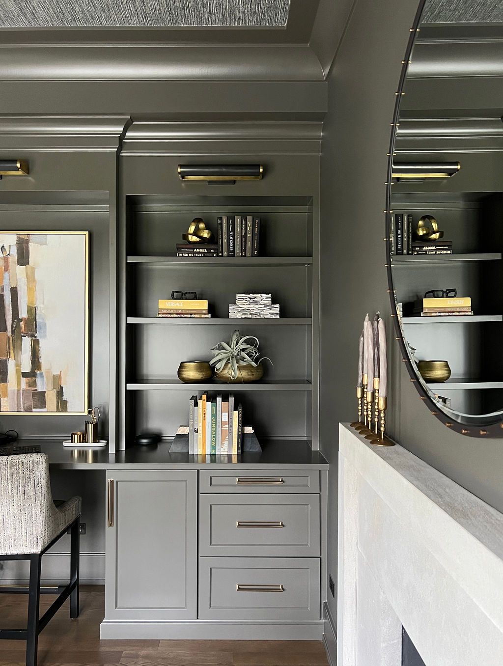 Built-in gray shelves and desk with gold accents, framed artwork, round mirror, and a fireplace.