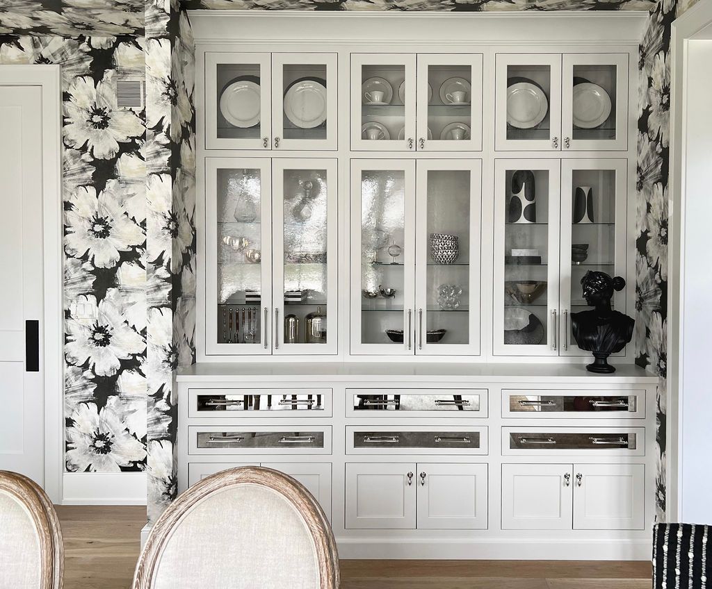 White built-in cabinet with glass-front doors, displaying dishes and decor, against black and white floral wallpaper.