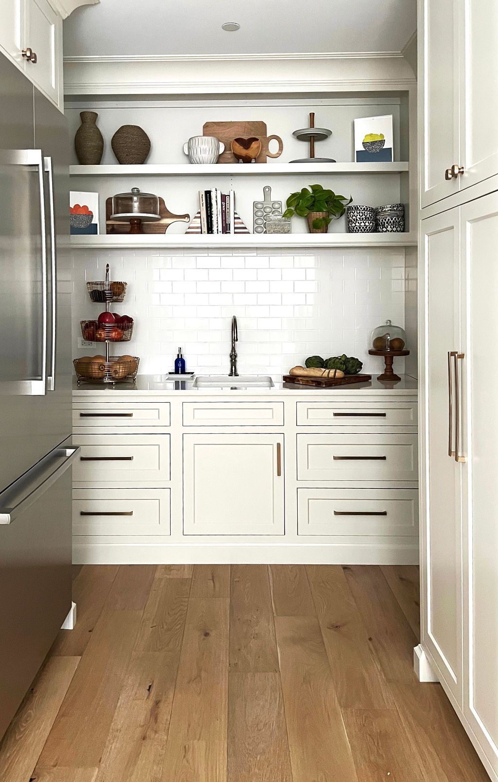 White kitchen pantry with shelves of decor and drawers. Light wood floor.