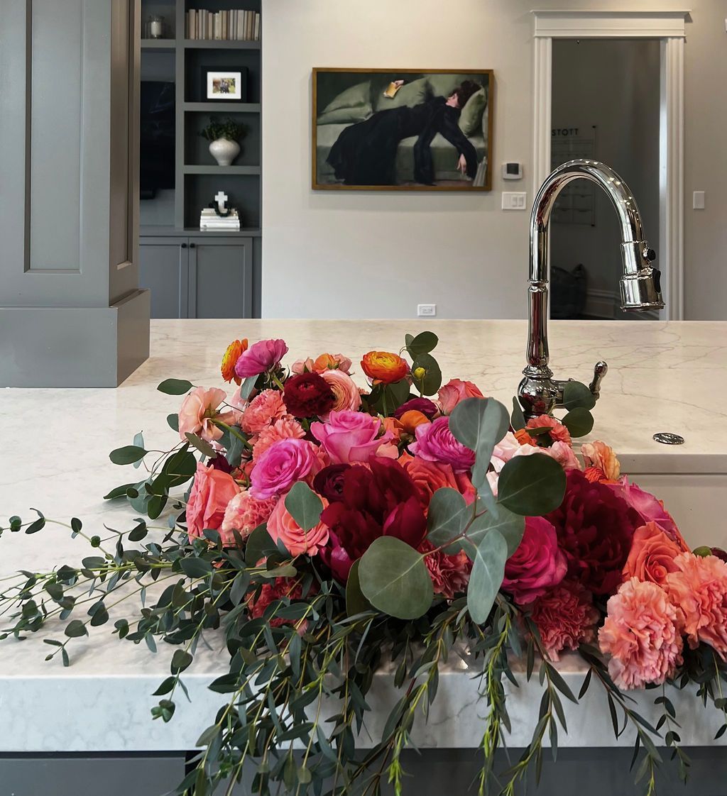 Close-up of vibrant flowers on a white countertop in a kitchen; a painting and cabinetry are in the background.