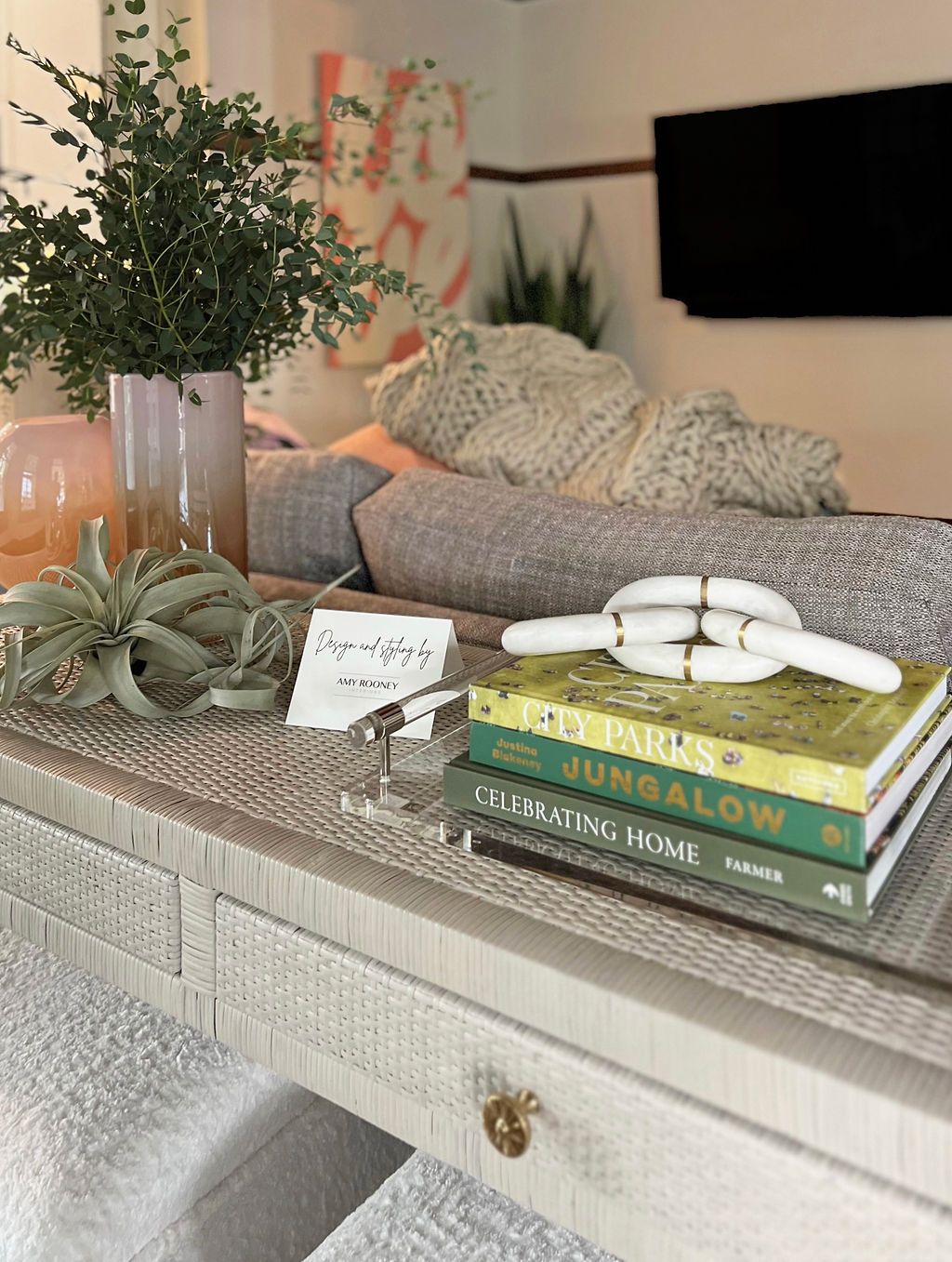 A console table decorated with books, plants, and a knot object. A couch and wall art are in the background.