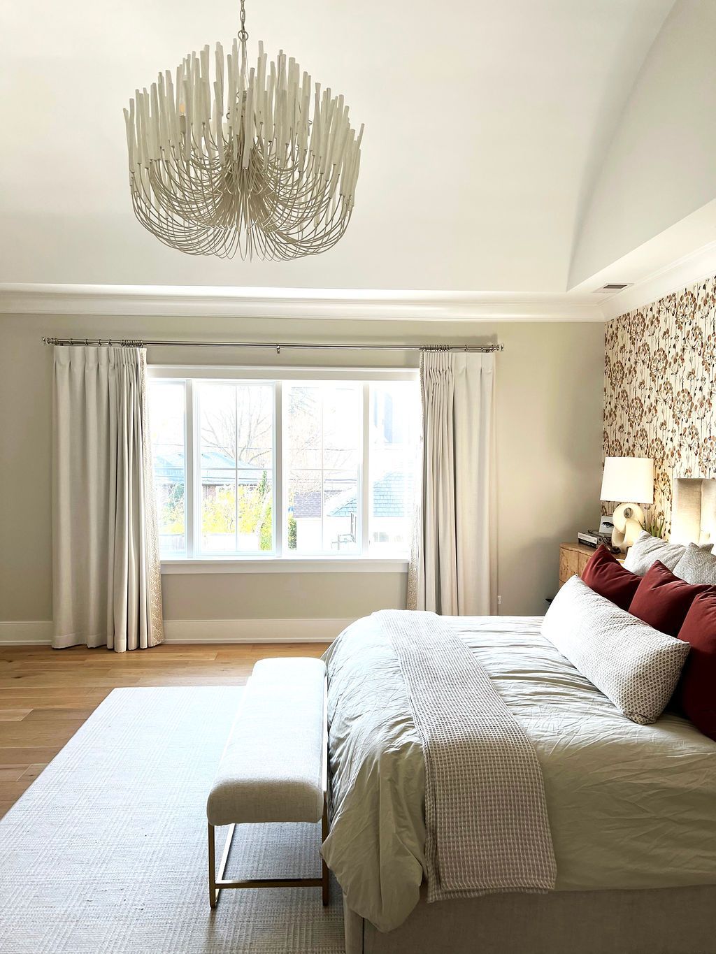 Bedroom with a white chandelier, large window, bed with red pillows, and neutral colored walls.