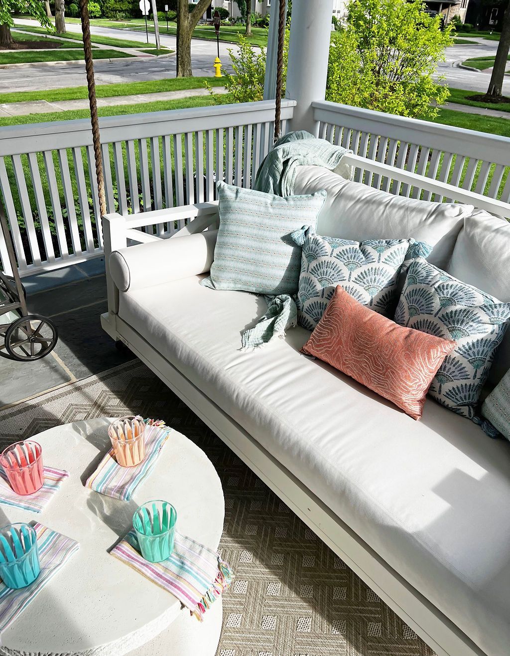 White porch swing with blue and coral pillows, a white table with colorful drinks, and a sunny porch.
