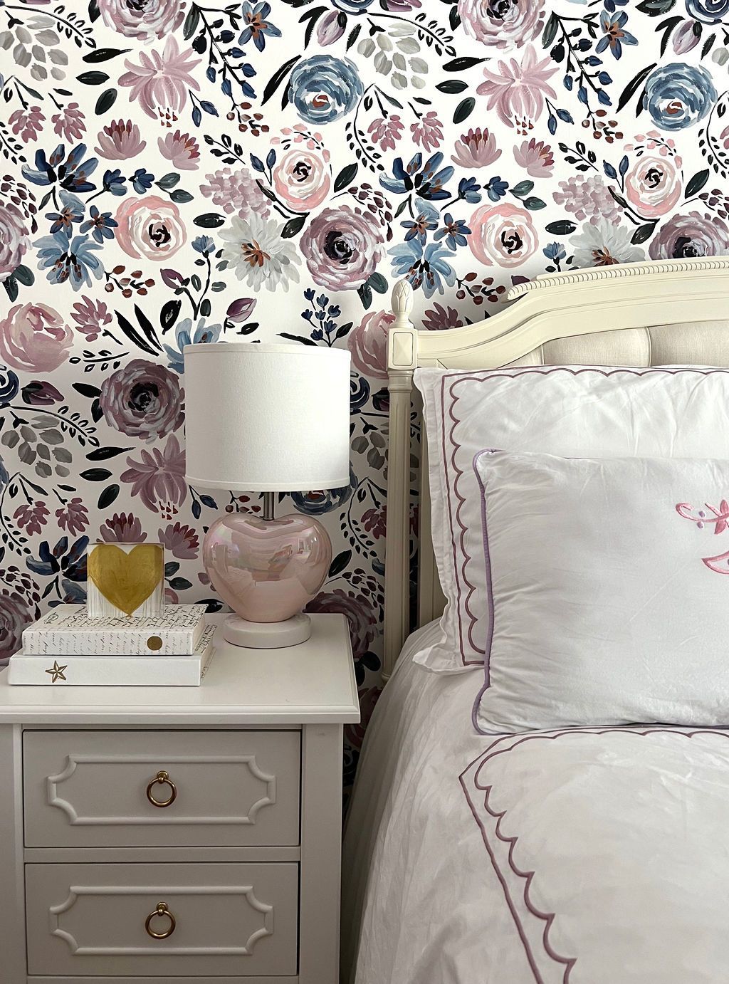 Bedroom with floral wallpaper, white nightstand, pink lamp, and bed with white linens and decorative pillow.
