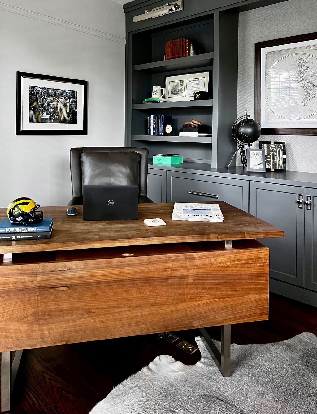 Wooden desk in a gray office with shelves, a chair, and a rug. Laptop and books are on the desk.