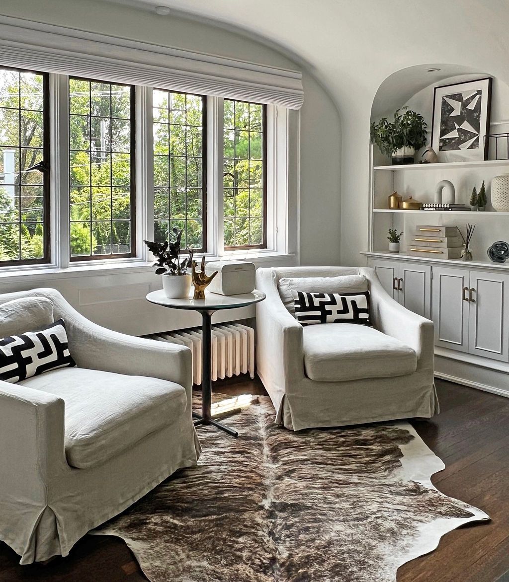 Cozy reading nook with white armchairs, cowhide rug, and built-in shelving next to arched windows.