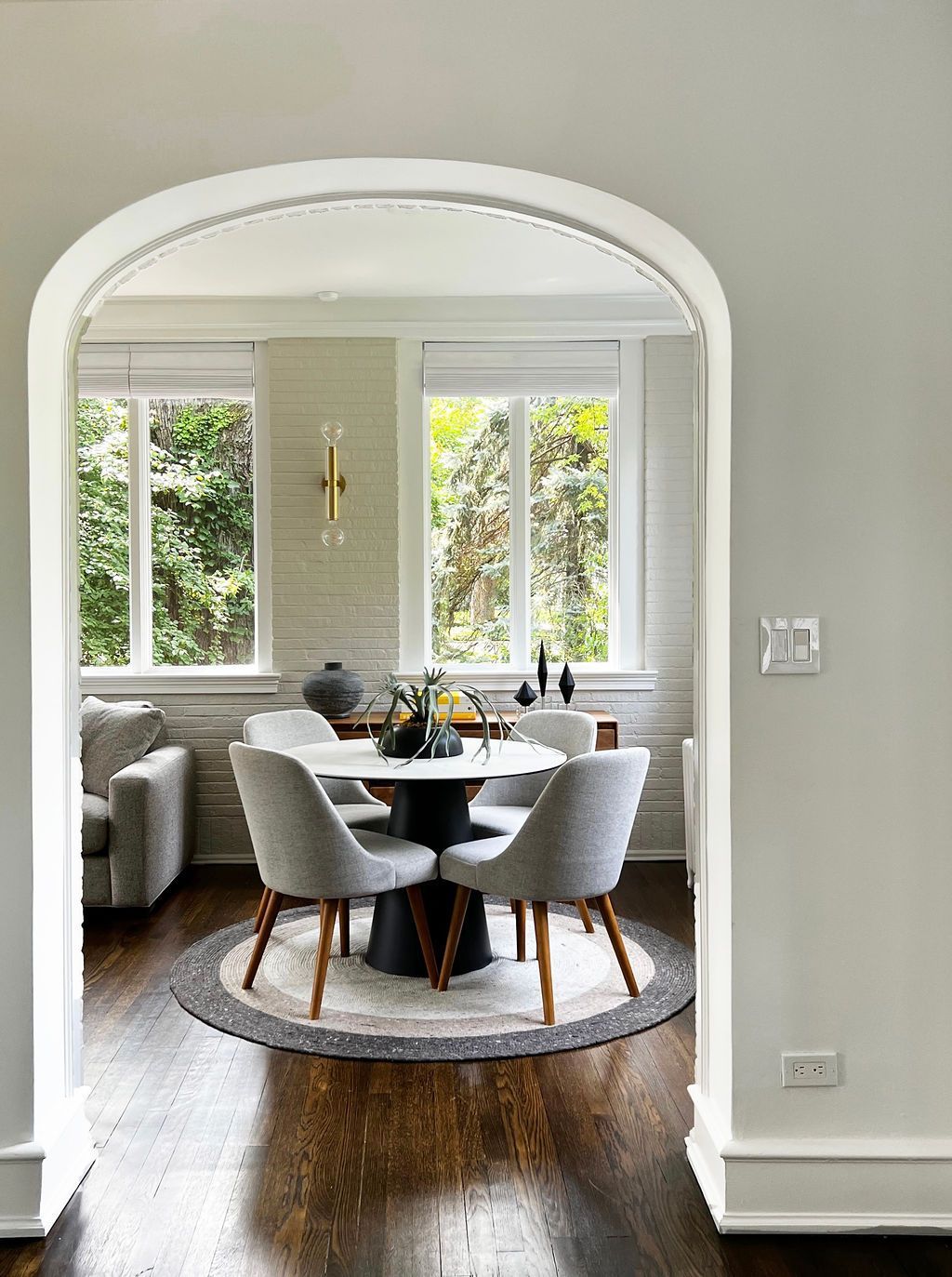 Dining room with a round table, chairs, and large windows. Archway frames the view.