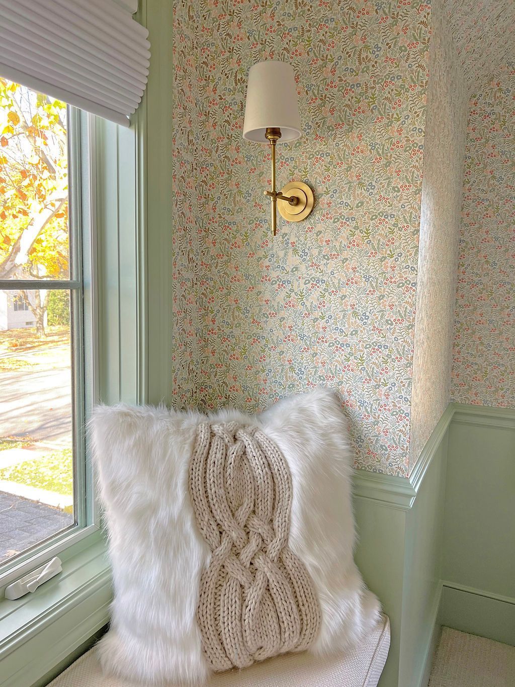 Cozy window seat with fur pillow, patterned wallpaper, and sconce. Light green trim, natural light.