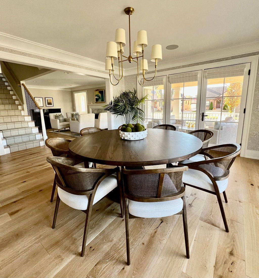 Round wooden dining table with six chairs, lit by a chandelier, in a bright room with hardwood floors.