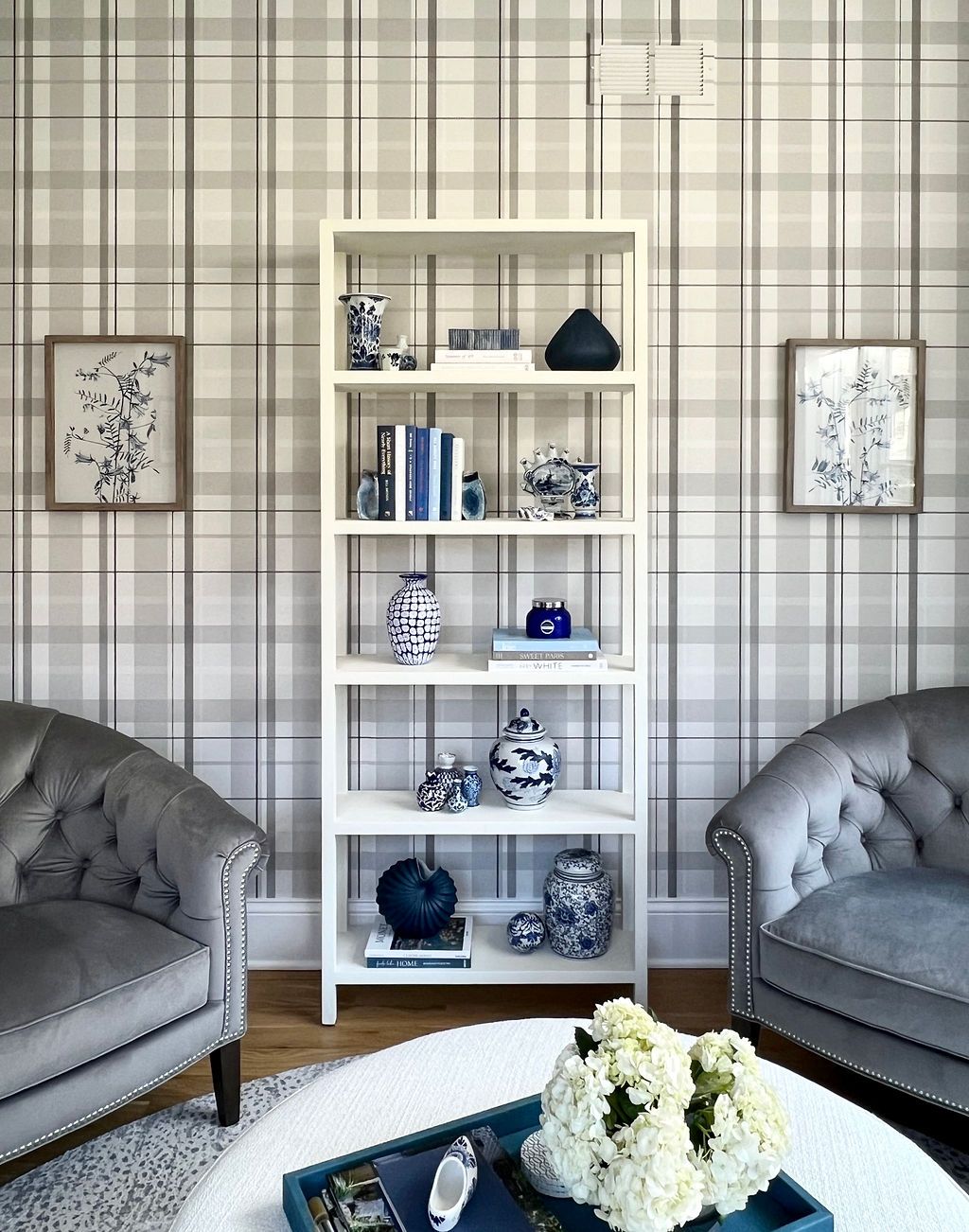 Cozy reading nook with gray plaid wallpaper, white bookshelf, blue decor, two gray armchairs, and a tray with flowers.