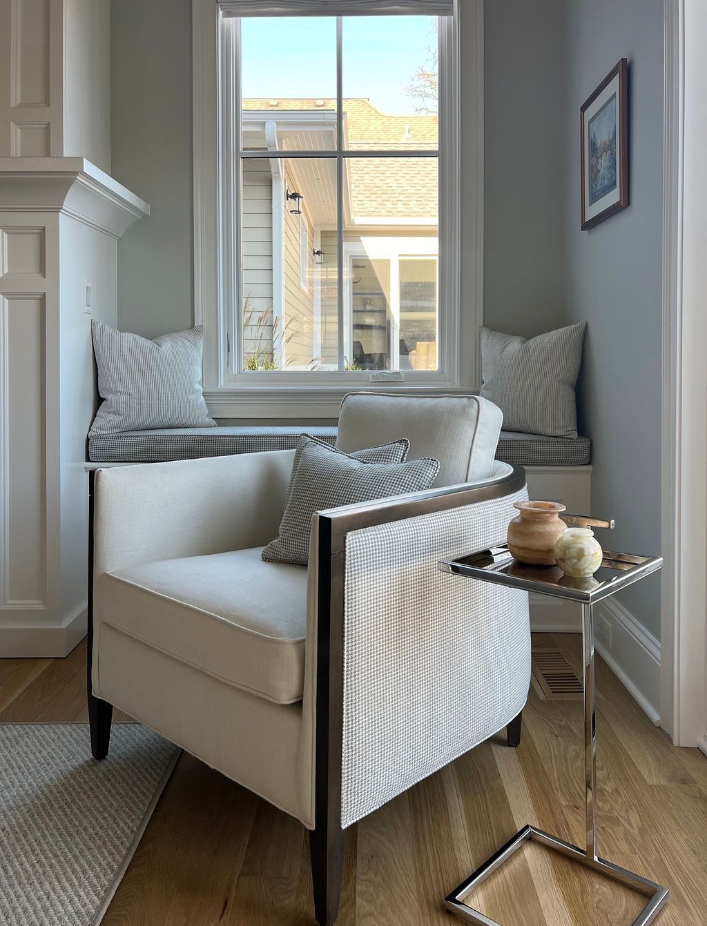 Cozy reading nook with white chair, window seat, and small side table. Soft blue walls, natural light.