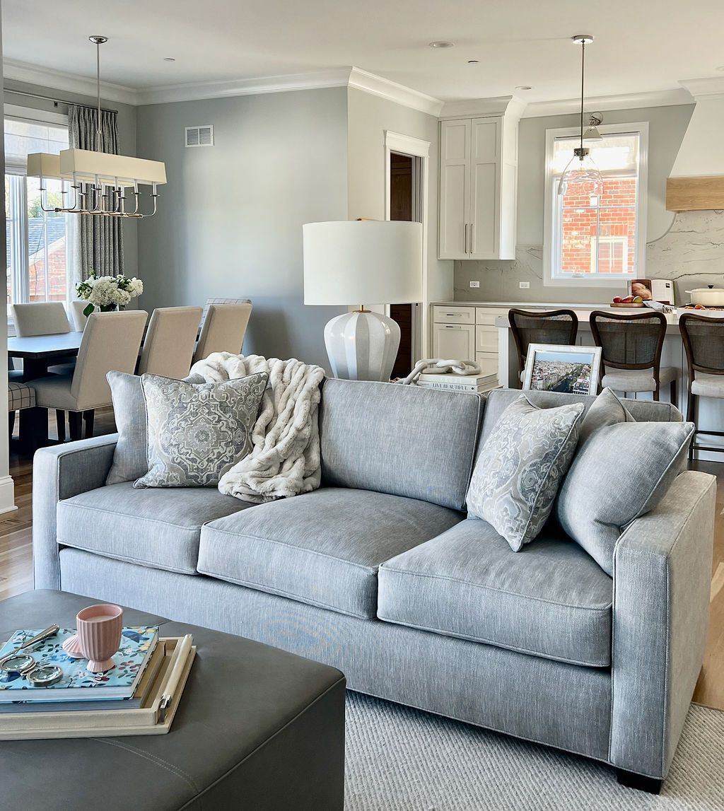 Gray sofa with throw pillows and blanket in a light-filled living room; dining area and kitchen visible.