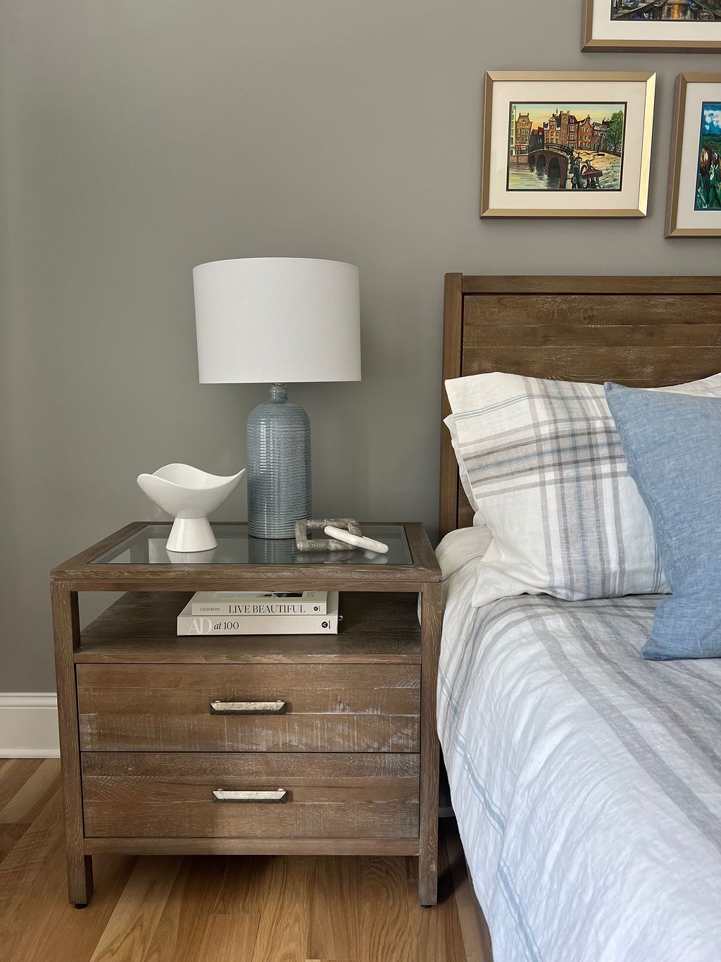Wooden nightstand with lamp, decor, and framed art beside a bed with blue and white bedding.