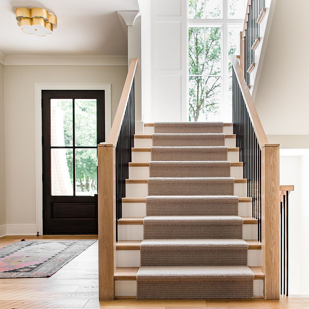 Staircase with light brown treads and a beige carpet runner, black railing. Entrance door.