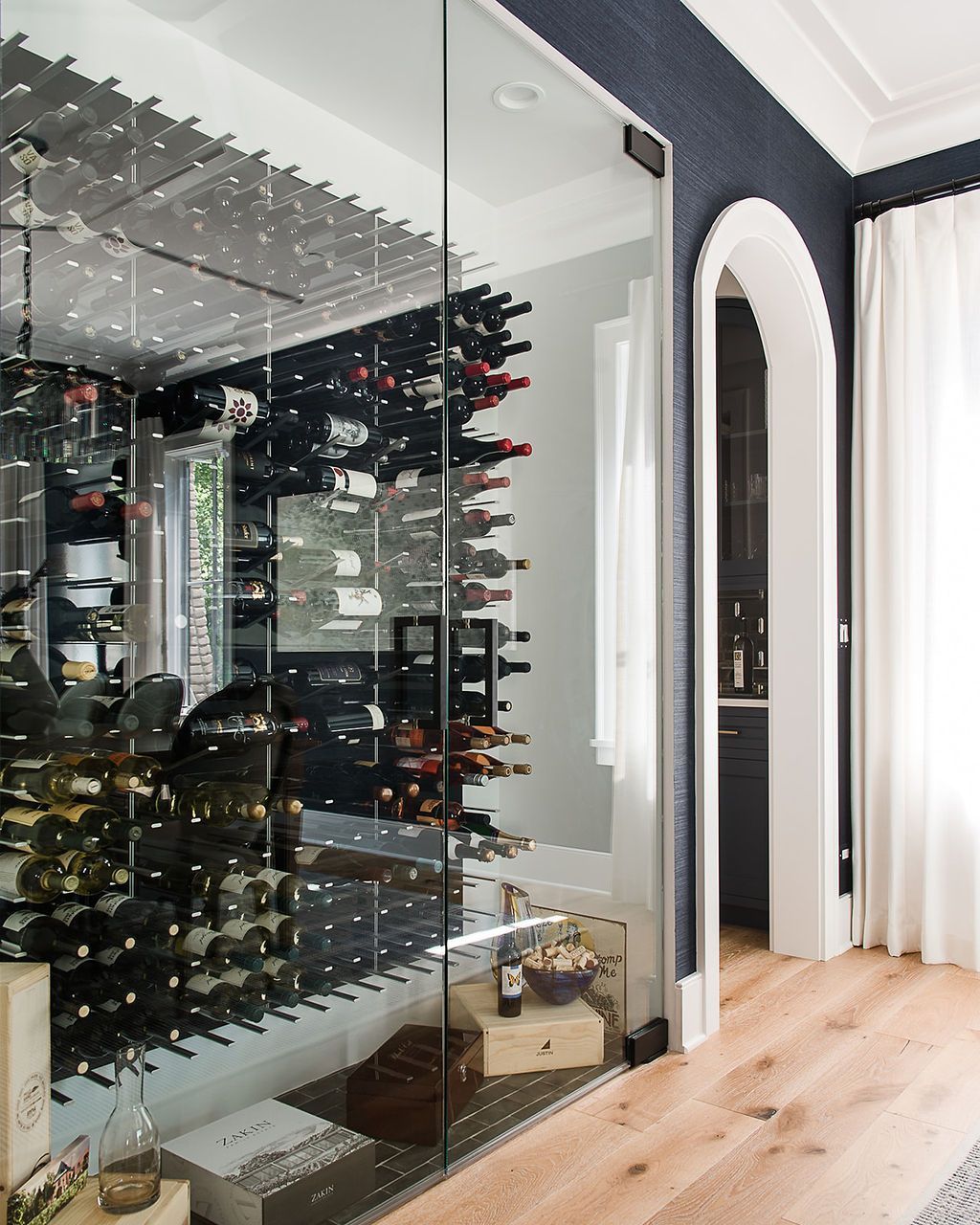 Wine cellar with glass door, showcasing numerous wine bottles on racks, with wooden floors and arched doorway.