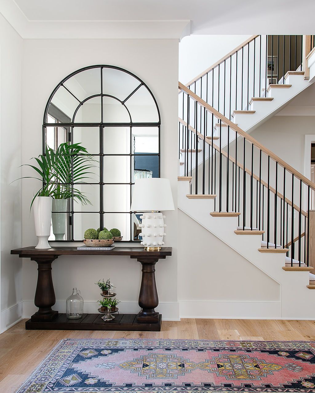 Entryway with arched mirror, console table, and staircase; floral rug on wooden floor.