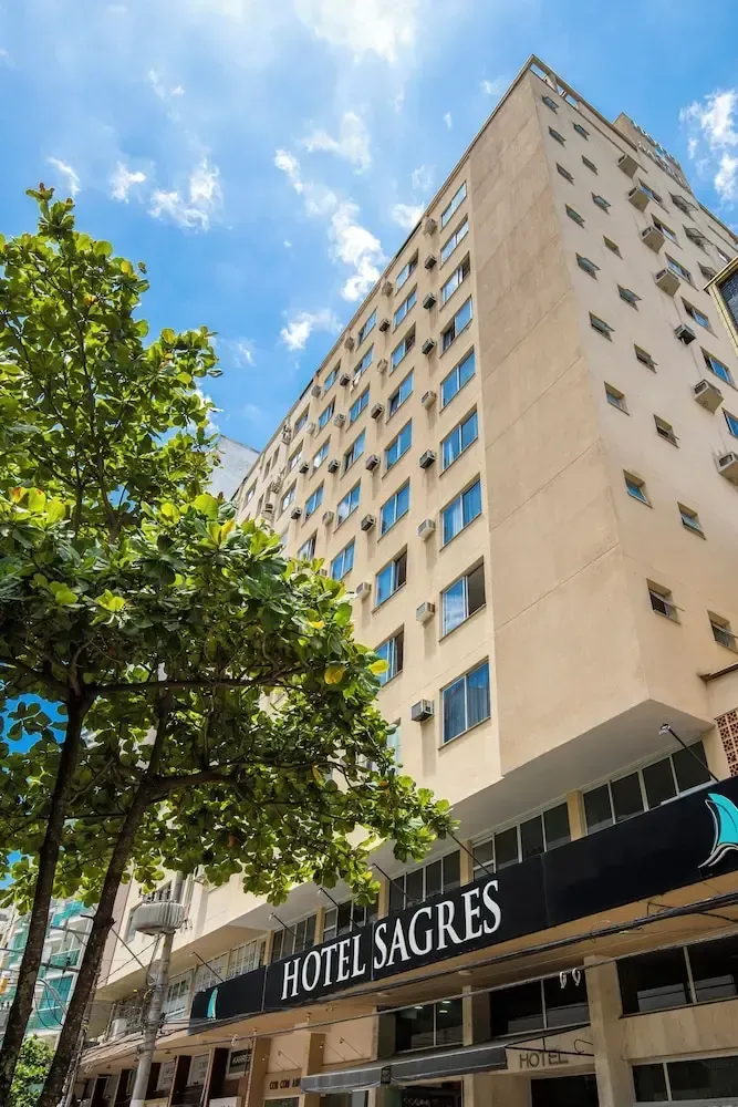 Edificio de hotel alto de color beige con ventanas azules y un árbol enfrente bajo un cielo azul brillante.