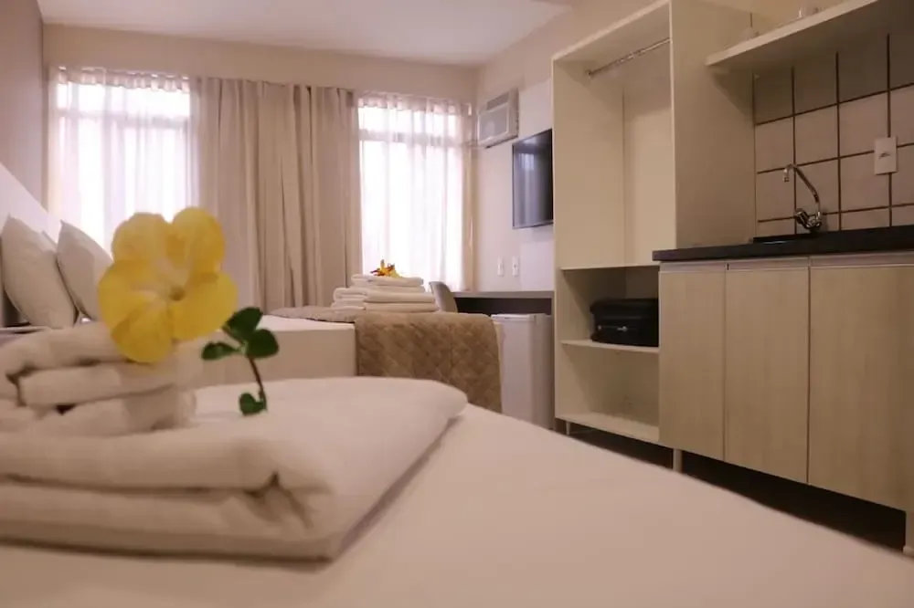 Habitación luminosa con sábanas blancas, una flor amarilla y una pequeña cocina junto a grandes ventanales.
