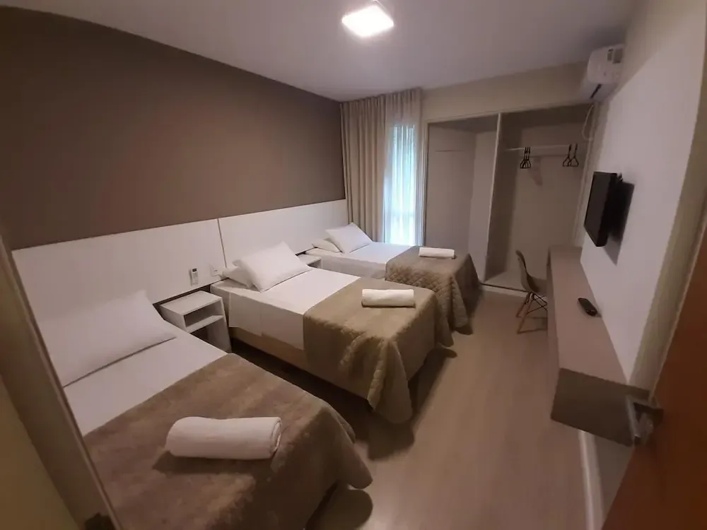 Habitación luminosa con sábanas blancas, una flor amarilla y una pequeña cocina junto a grandes ventanales.