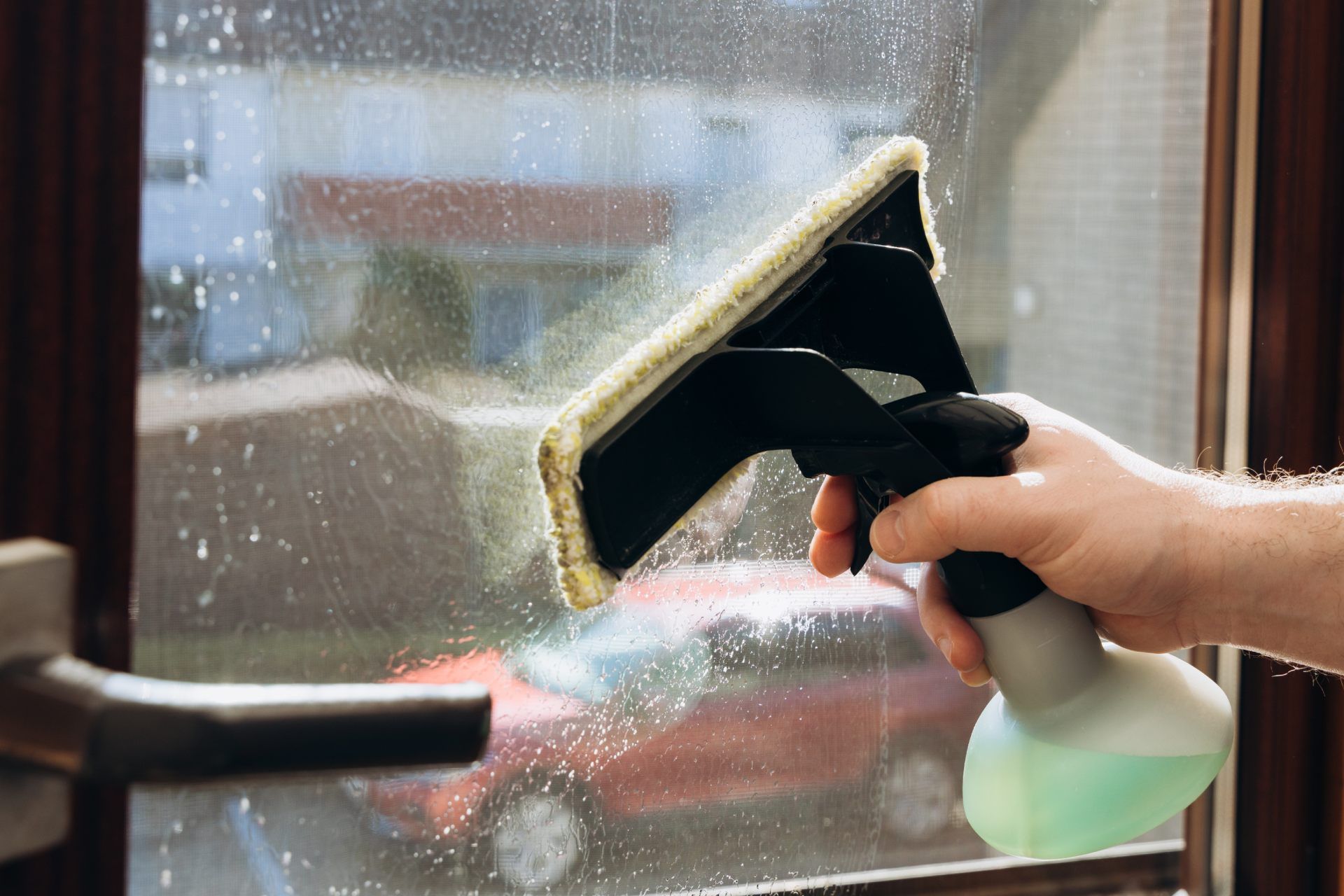 A hand uses a squeegee tool with a cleaning pad to wipe soap and water off a window, with a car visible outside.