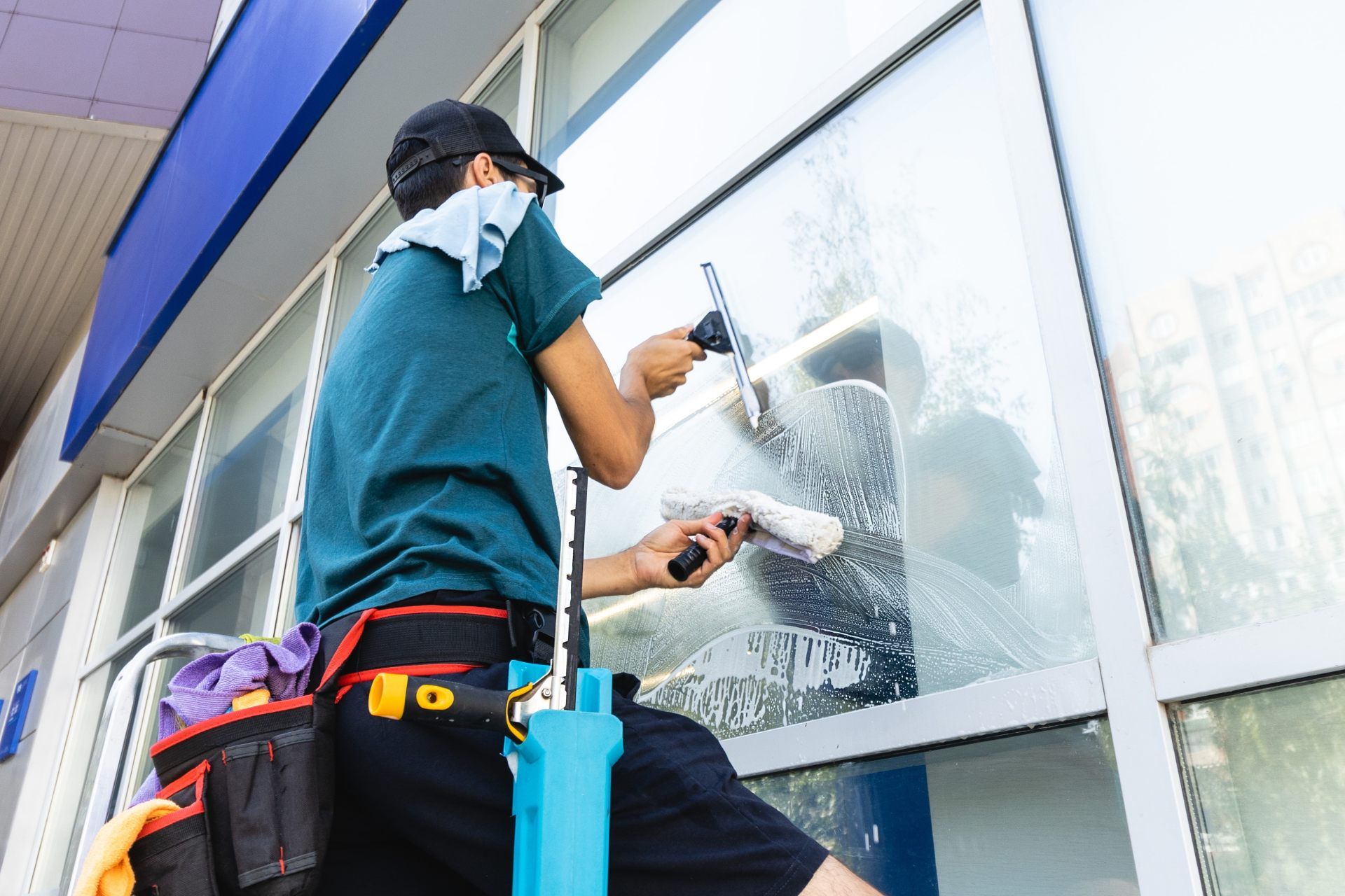 A worker in a teal shirt uses a squeegee and cloth to clean a large glass storefront window.