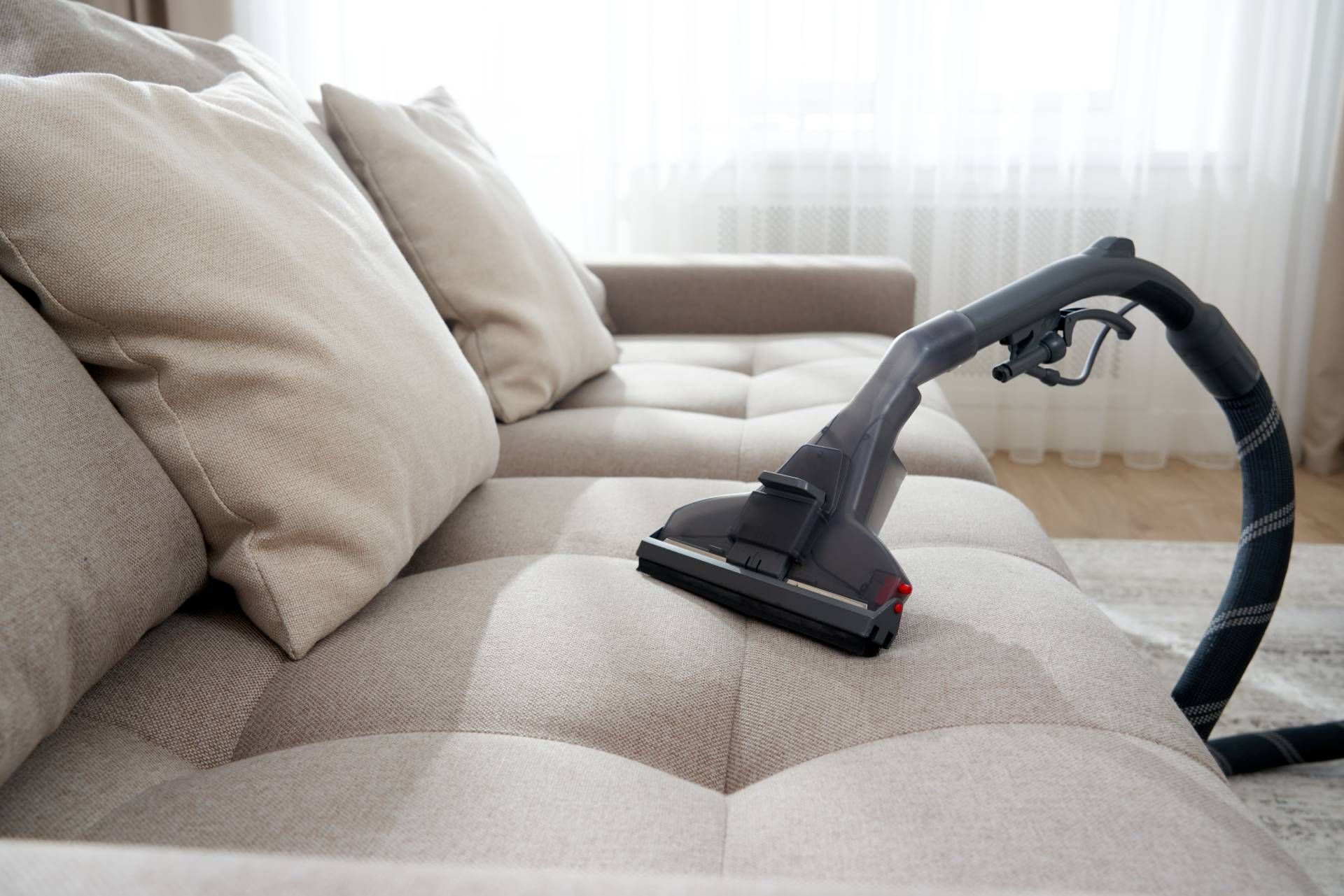 A vacuum cleaner nozzle cleans a light-colored, tufted fabric sofa near a bright window.