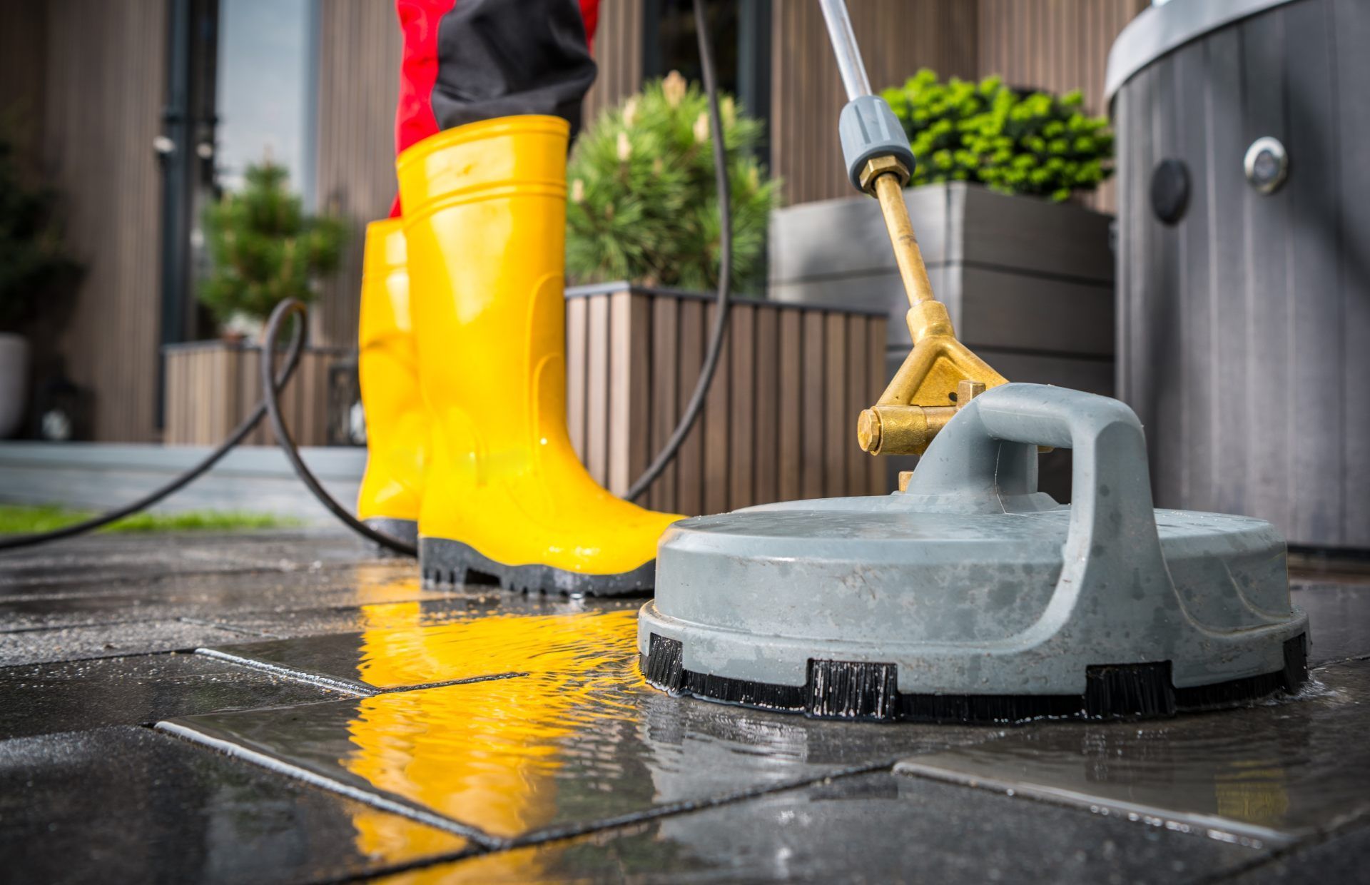 A person in bright yellow rubber boots uses a circular surface cleaner to wash a dark stone patio.