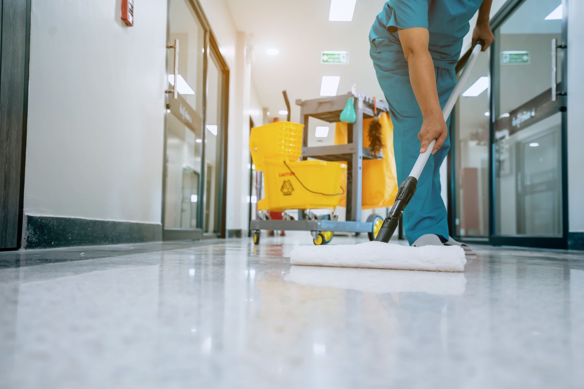 A person in teal scrubs mops a polished floor in a bright hospital hallway with a cleaning cart nearby.