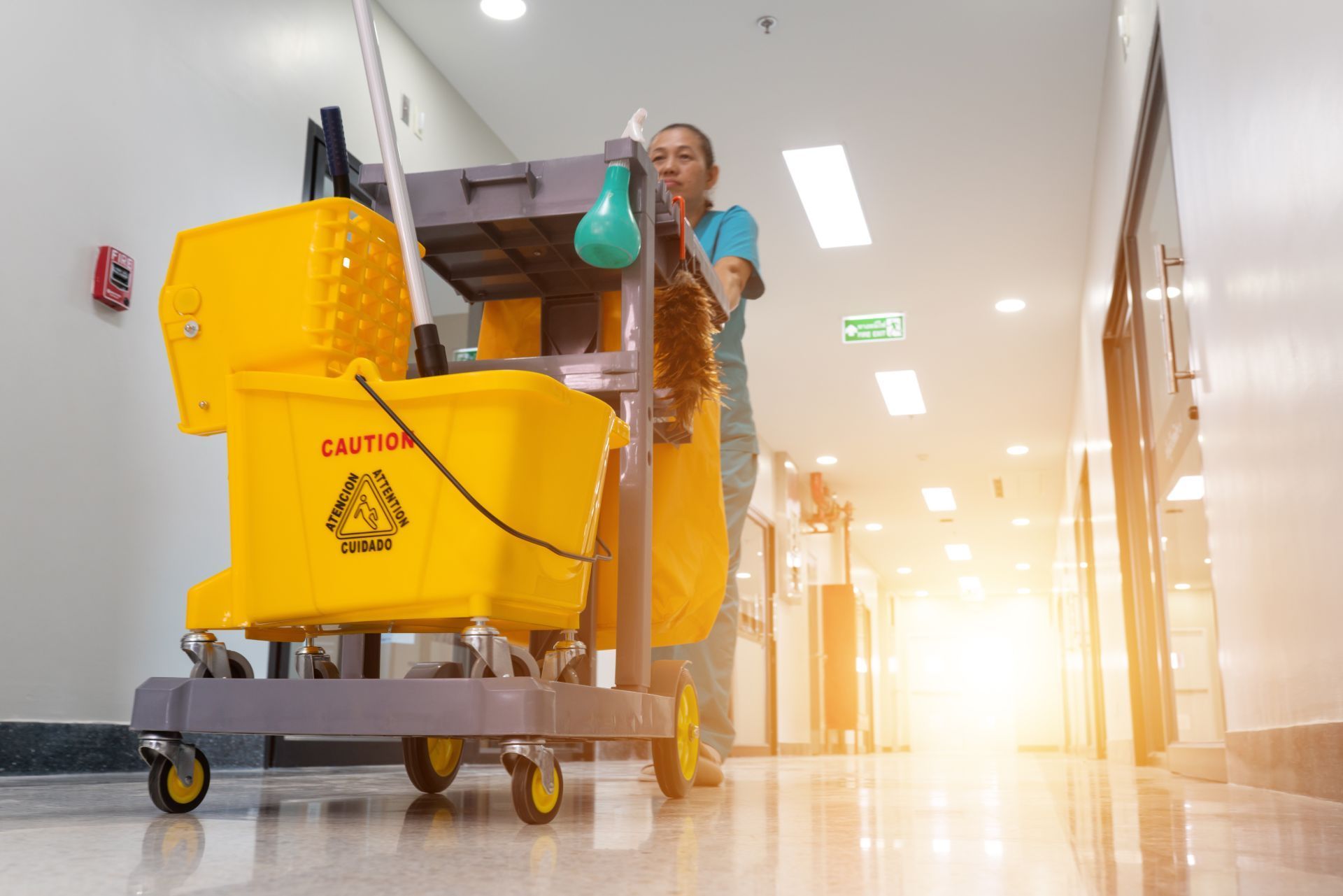 A person pushes a yellow janitorial mop cart down a bright, sunlit hospital or office corridor.